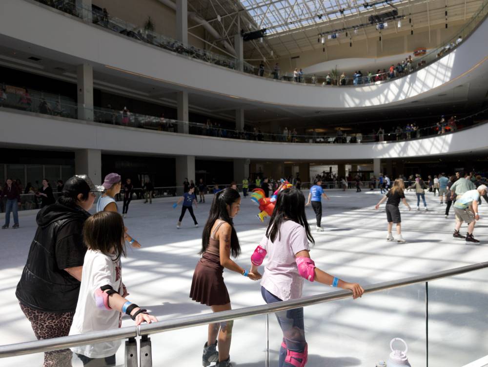 The ice rink, full of people skating, including a person in a colorful chicken outfit slightly obscured in the middle of the rink. There are a lot of people overlooking the rink on balconies surrounding it.