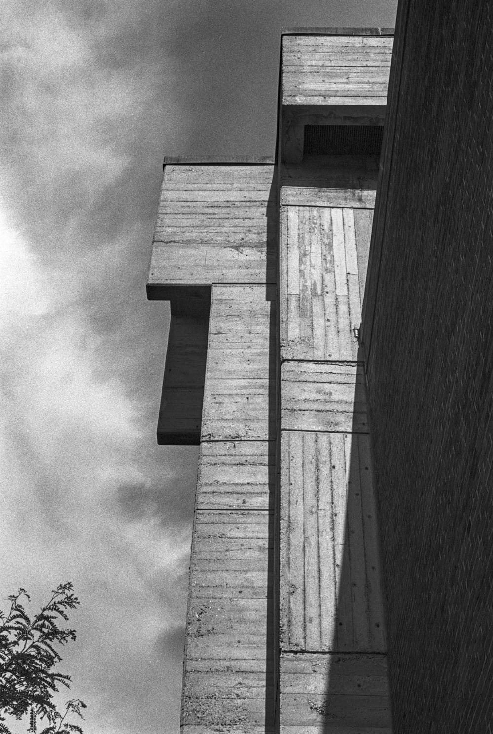 Tight upward view of a brutalist building corner. Board-formed concrete slabs, their wood-grain shuttering marks clearly visible, step outward in a staggered cascade against a hazy sky. To the right, a brick wall recedes into shadow. A single tree branch enters the lower left of the frame.