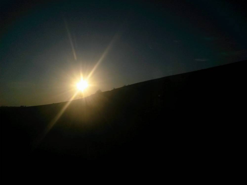 A bright sun sits low on the horizon, creating a prominent starburst lens flare against a dark twilight sky. Below it, a completely silhouetted hillside slopes upwards from the left to the right side of the frame. There appear to be small bumps along the ridge that are possibly distant trees or shrubs, though the deep shadow makes it uncertain... my eyes fe02eabd-7035-4b0f-a1f1-3f57bdd51ee0