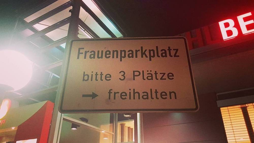 A German sign reading "Frauenparkplatz, bitte 3 Plätze freihalten" (women's parking, please keep 3 spaces free) outside a building at night.