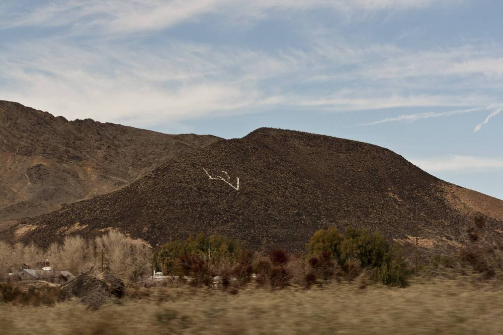 A rocky desert hillside with a large letter shape formed from stones is seen from a moving car.