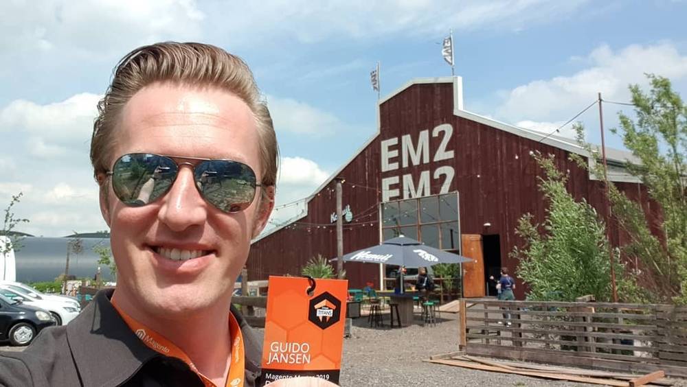 A man wearing sunglasses takes a selfie with a MageTitans badge in front of the EM2 barn building.