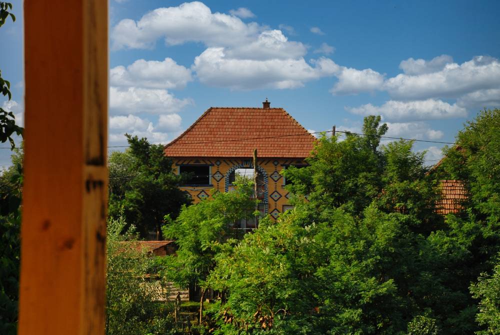 Paisaje rural con una casa de estilo tradicional decorada con llamativos motivos geométricos pintados en la fachada, en tonos ocres, azules y blancos, con formas de rombos y estrellas. El tejado a cuatro aguas está cubierto de tejas de terracota rojiza y rematado por una chimenea. La vivienda se alza tras una densa masa de árboles y arbustos de follaje verde, que ocultan parcialmente su planta baja. A la izquierda, un poste de madera de color anaranjado aparece desenfocado en primer plano, enmarcando la escena. El cielo es de un azul intenso salpicado de nubes blancas y algodonosas, y se distinguen cables eléctricos cruzando la imagen.