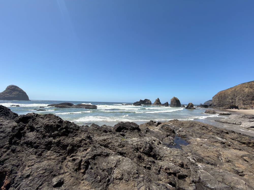 Ocean shoreline viewed from a rocky outcrop. Several sea stacks stand offshore while waves roll toward the beach. Dark volcanic-looking rocks fill the foreground under a bright blue sky.