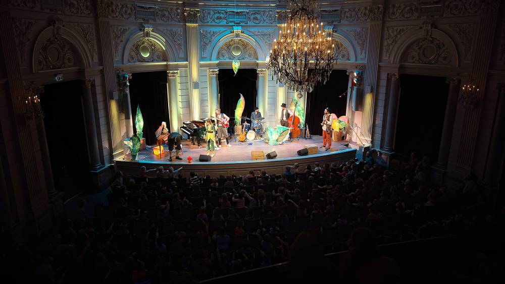 A band performs on stage in an ornate theatre, seen from a balcony above the crowd.