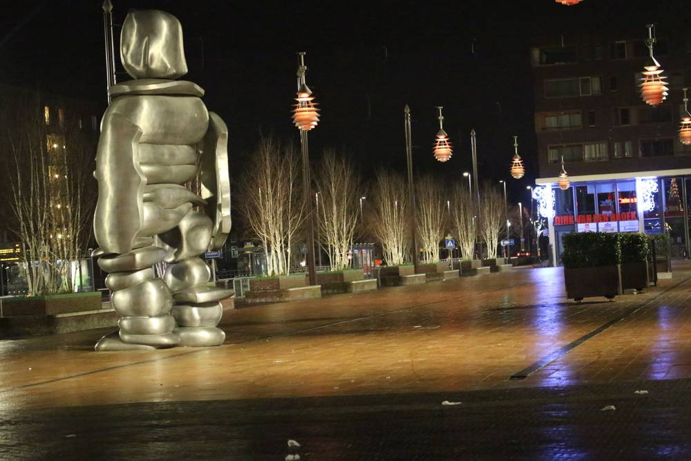 A tall silver sculpture of a robed figure stands in a lit plaza at night.