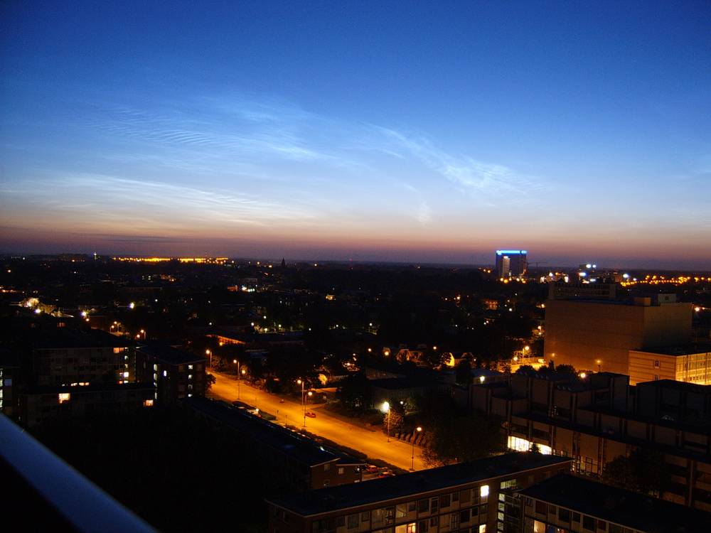 Wispy noctilucent clouds glow blue against the dark sky above the Utrecht skyline at night.