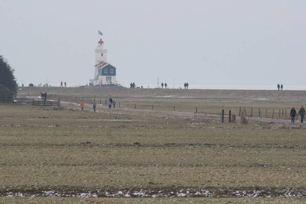 A distant lighthouse on Marken seen across a snow-dusted field with people walking along a path.