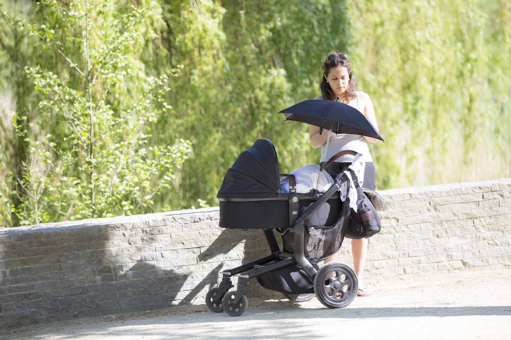 A woman pushes a pram with an umbrella along a shaded park path.