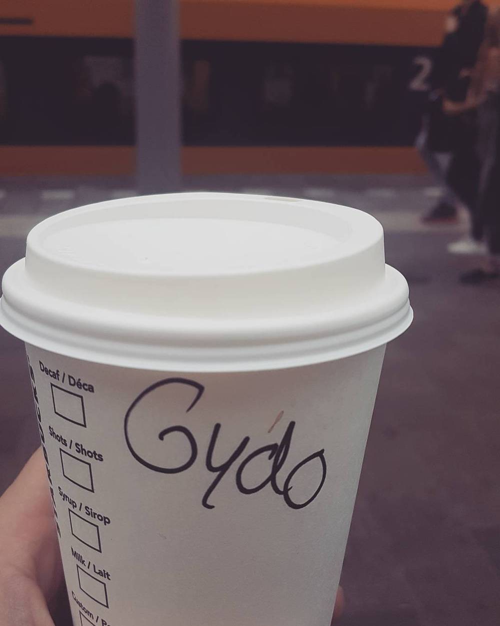 A takeaway coffee cup with the name "Gydo" misspelled on it, held on a train platform.
