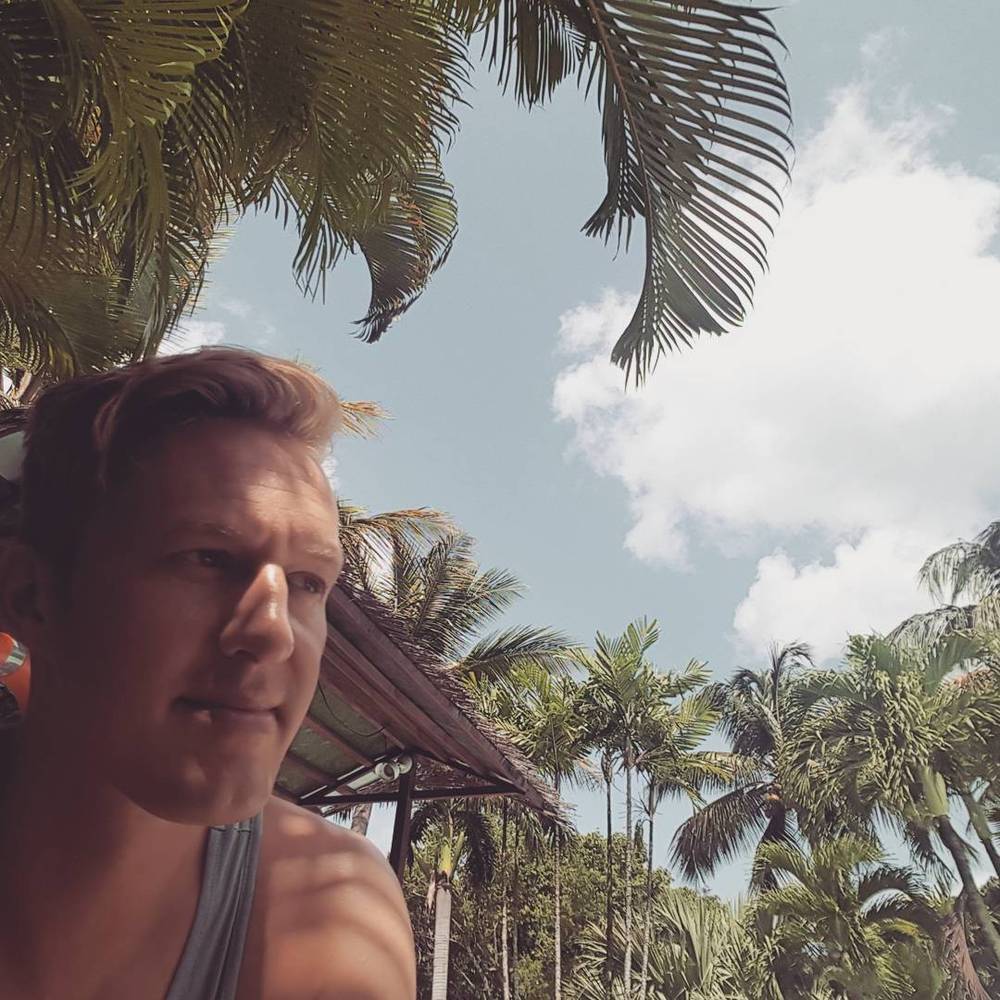 Selfie of a man with palm trees and a thatched roof in the background.