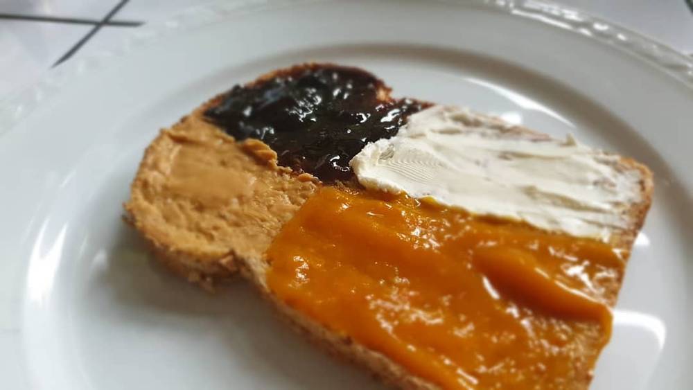 A slice of bread on a plate is spread with three different toppings: chocolate, peanut butter and marmalade.