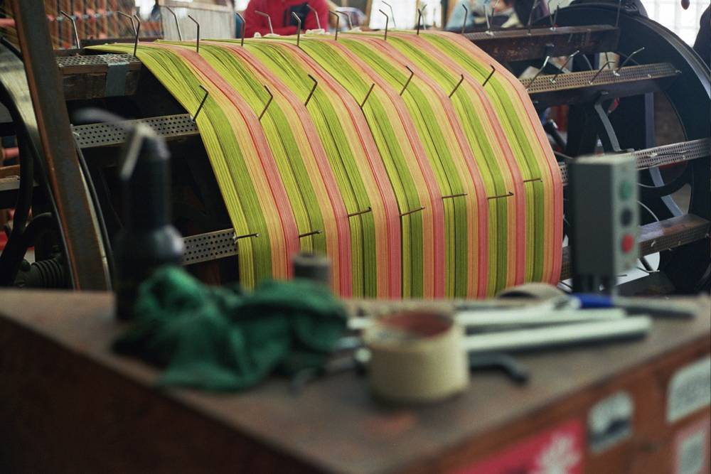 A curved sheet of warp threads in alternating stripes of chartreuse, coral, and pink stretched over a sectional warping drum, with scissors, tape, and a green cloth on the workbench in the foreground.