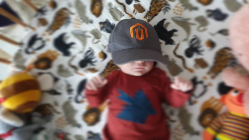 Blurred photo of a toddler wearing a Magento baseball cap and red sweater against a patterned background.