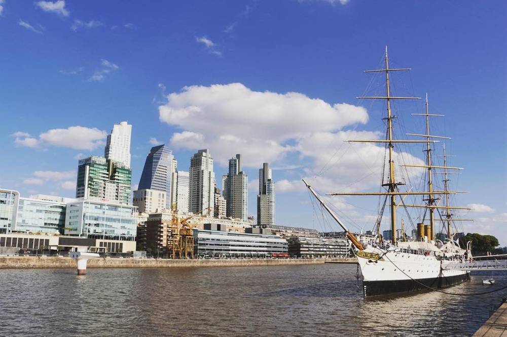 A tall-masted sailing ship moored in a harbor surrounded by modern skyscrapers.