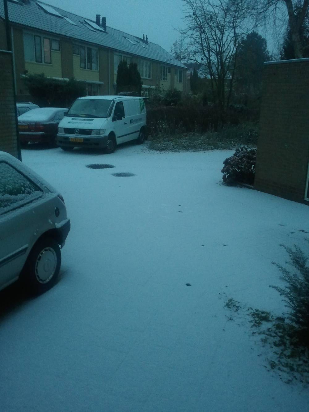 Snow dusts a quiet residential street lined with parked cars and vans.