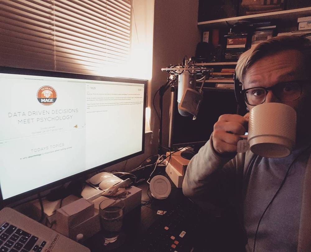 A man drinking coffee at his home desk, with a screen showing a presentation titled "Data Driven Decisions Meet Psychology."