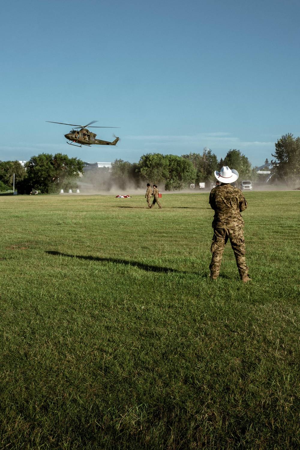A helicopter lands in a grassy field kicking up dust, as a person in military fatigues and a white cowboy hat looks on