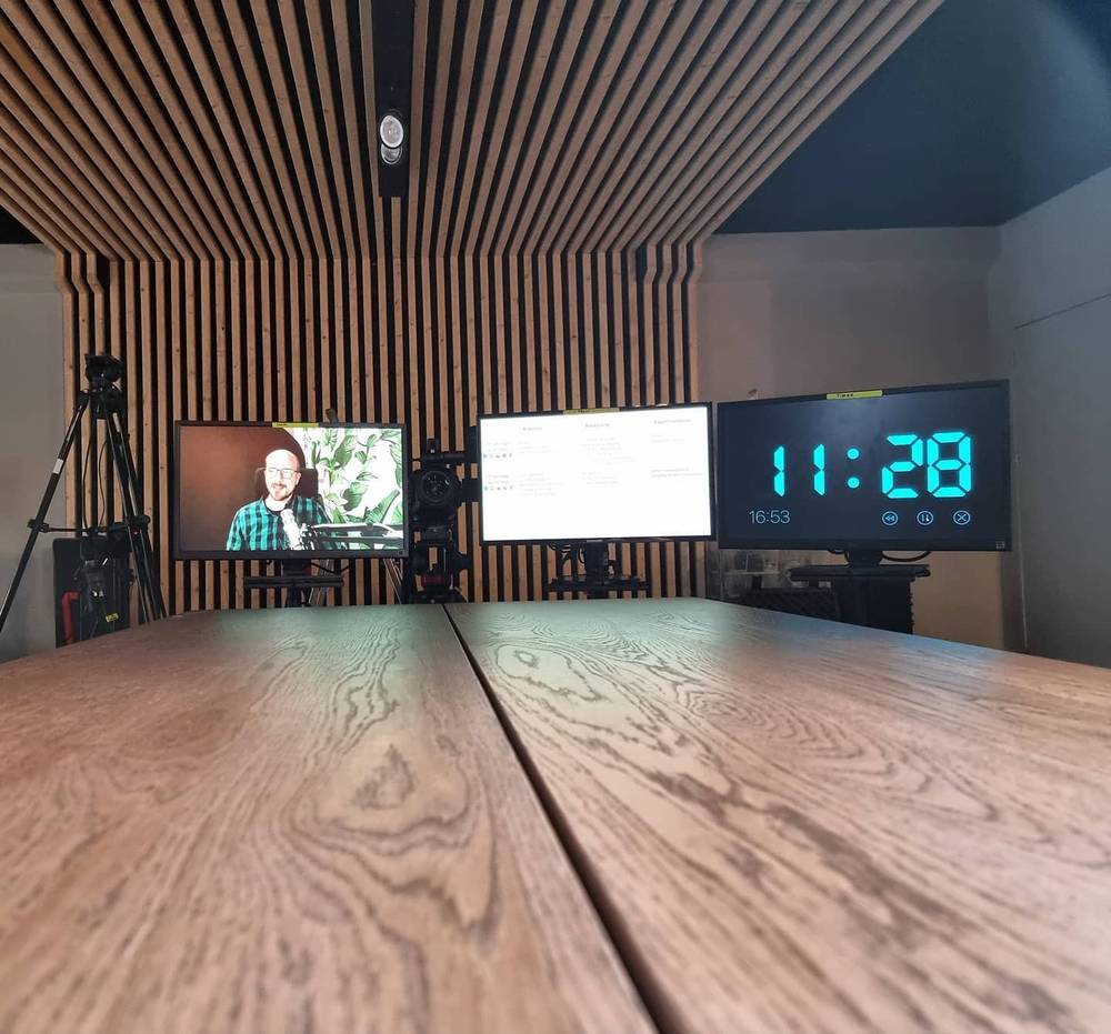 A home recording studio with slatted wood walls, three monitors on a desk, one displaying the time 11:28.