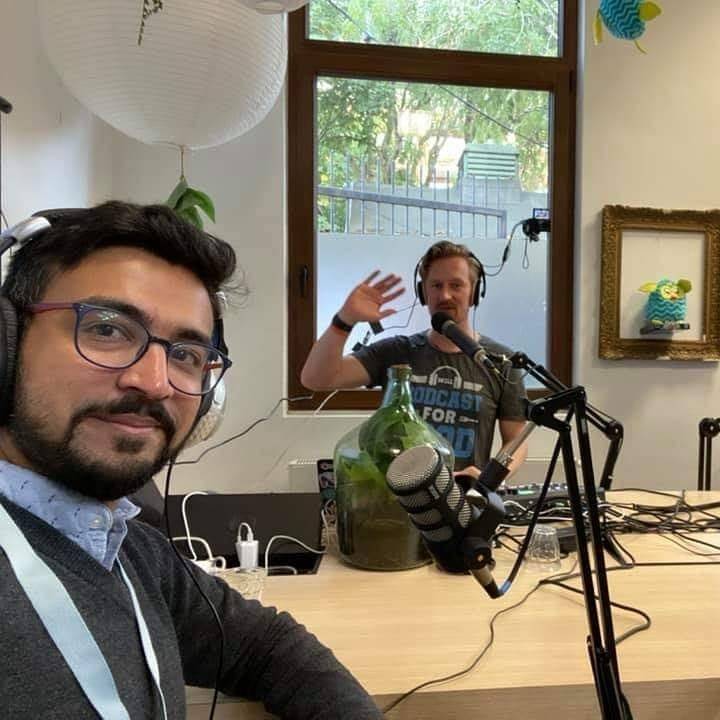 Two men record a podcast in a room, one waving, decorative items on the wall.