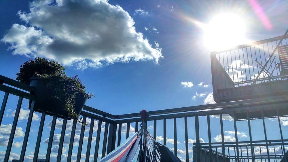 A balcony with a plant box and hammock, seen against a bright sky with clouds and sun flare.