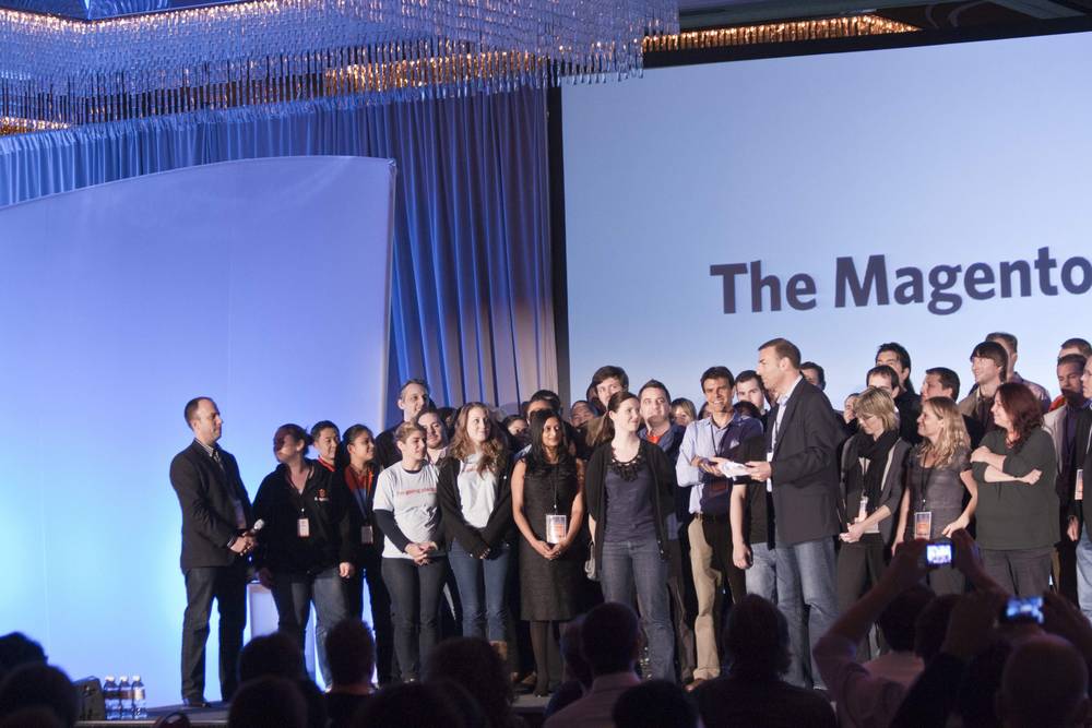 A large group poses on stage as a speaker addresses them beneath 'The Magento Family' screen.