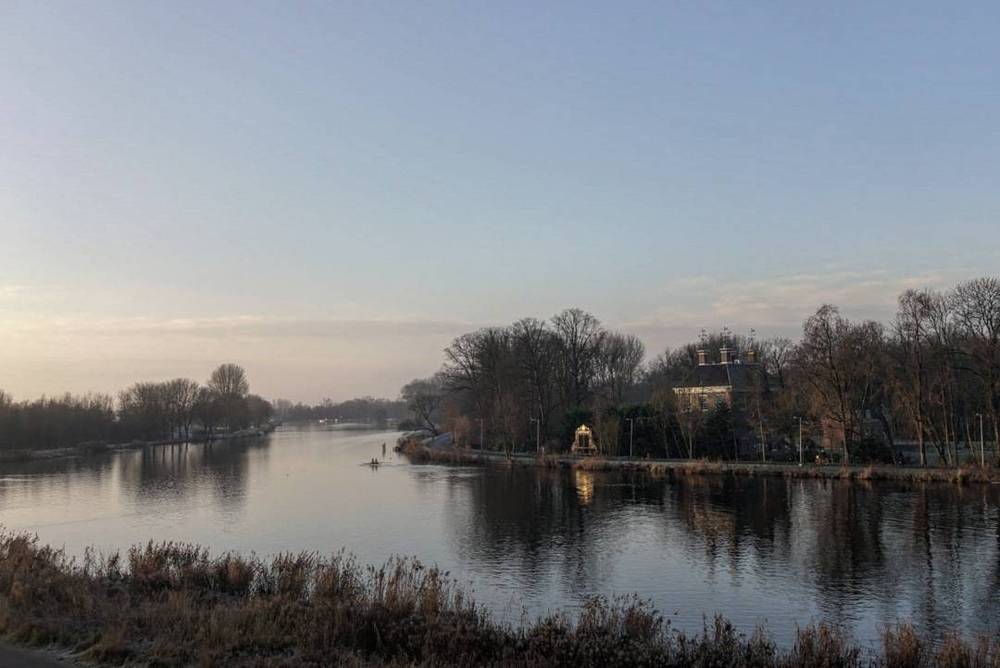 A calm river bordered by bare trees and grass at dusk.