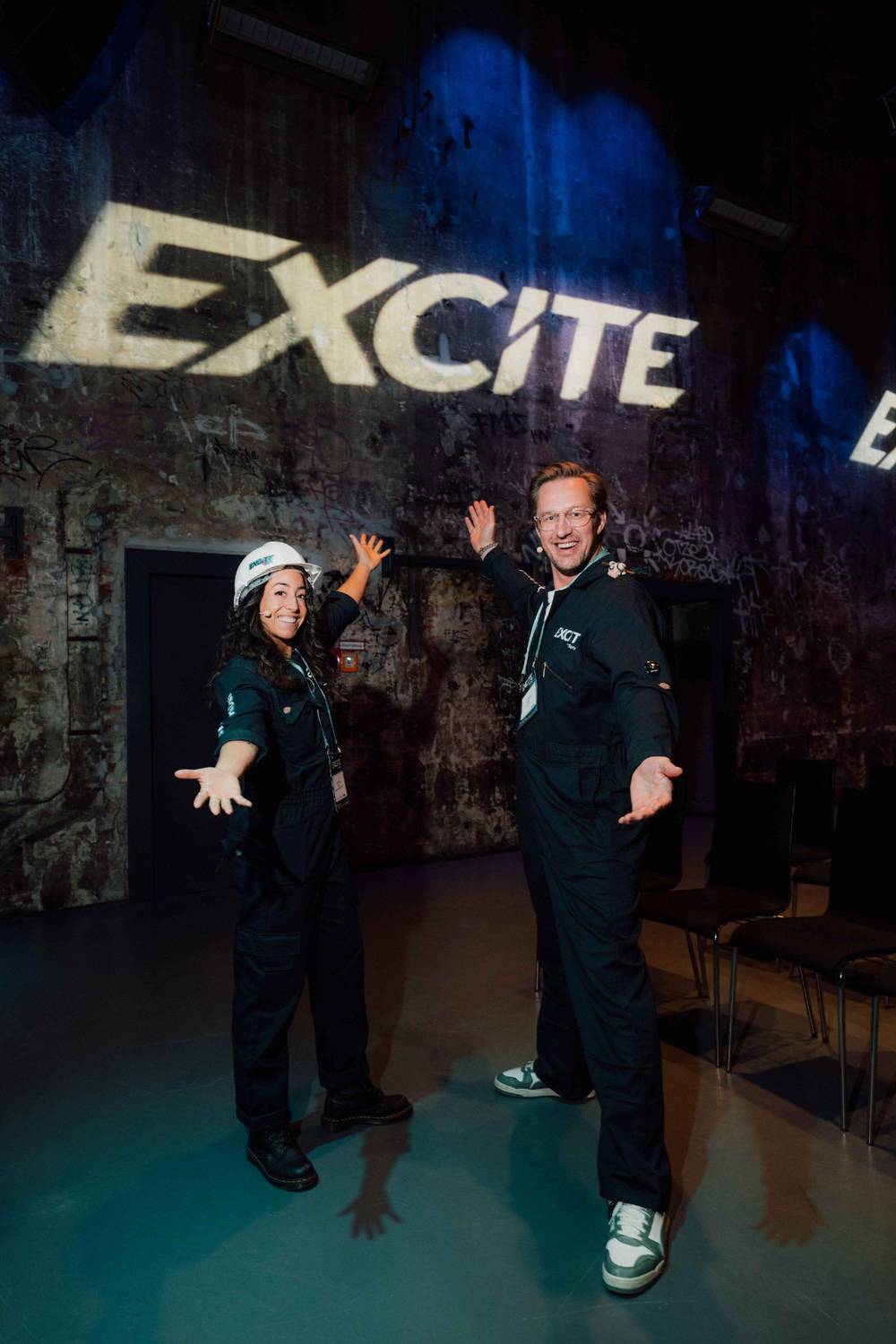 Photo of Cristina and Guido wearing their EXCITE coveralls, pointing at the EXCITE logo on the wall
