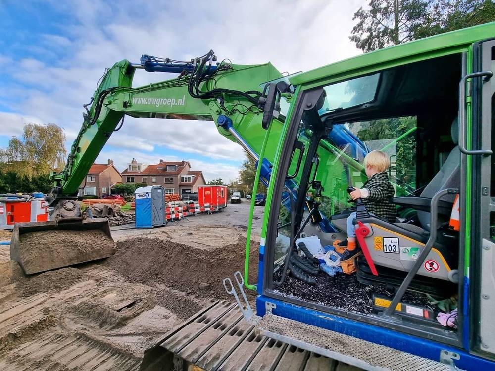 A green excavator at a road construction site, with a child sitting in the driver's cab.