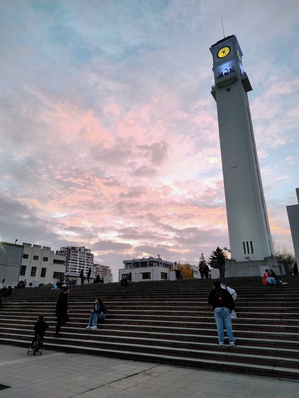 Vista desde una esquina del foro de la Universidad de Concepción hacia los escalones y el Campanil. En el fondo alcanza a aparecer la Facultad de Derecho, mientras las nubes stratocumulus reflejan el sol en un tono más rosado, junto al blanco y el celeste del cielo.