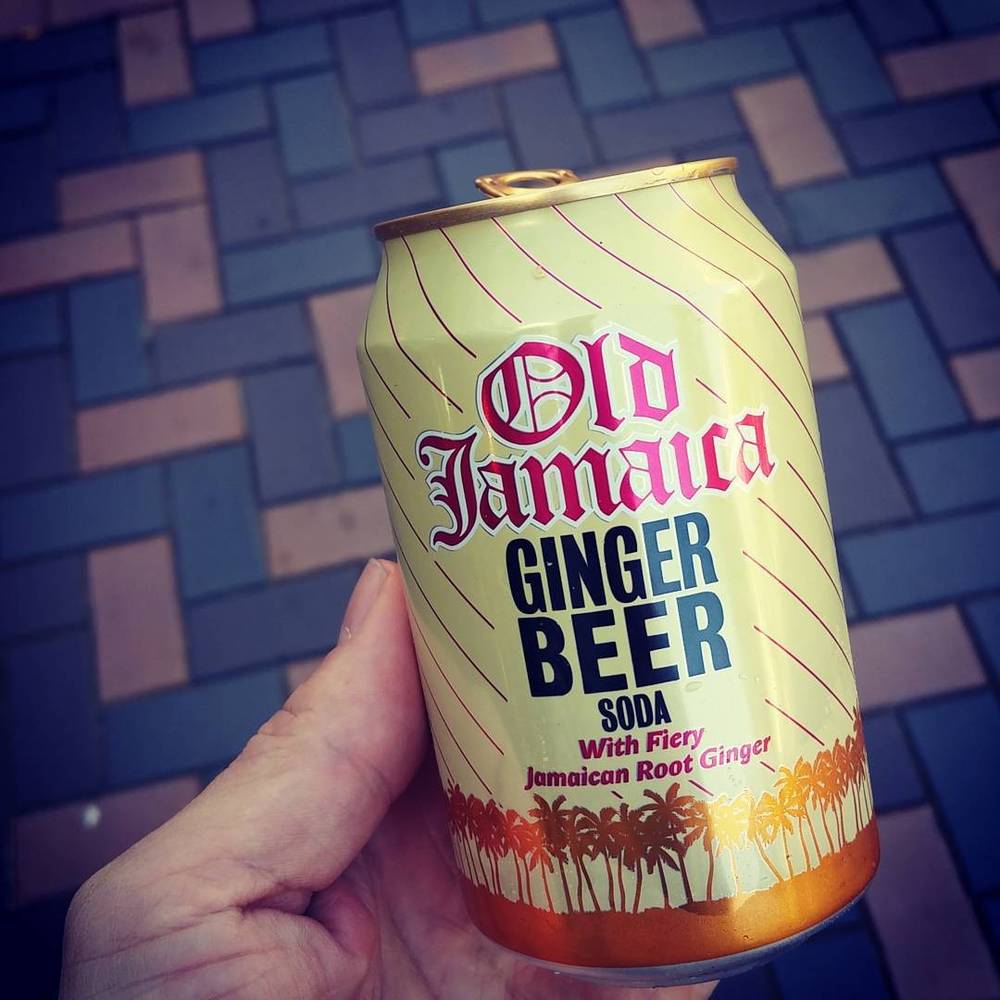 A hand holding a can of Old Jamaica ginger beer soda against a tiled wall.