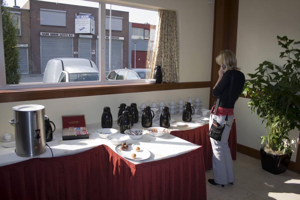 A woman looks out a hotel window past a breakfast buffet table set with coffee urns and pastries.