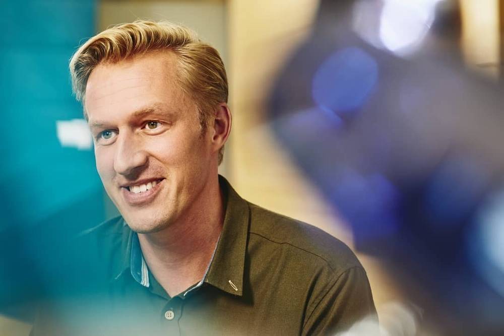 A close-up portrait of a smiling man during a video interview, blue lighting.