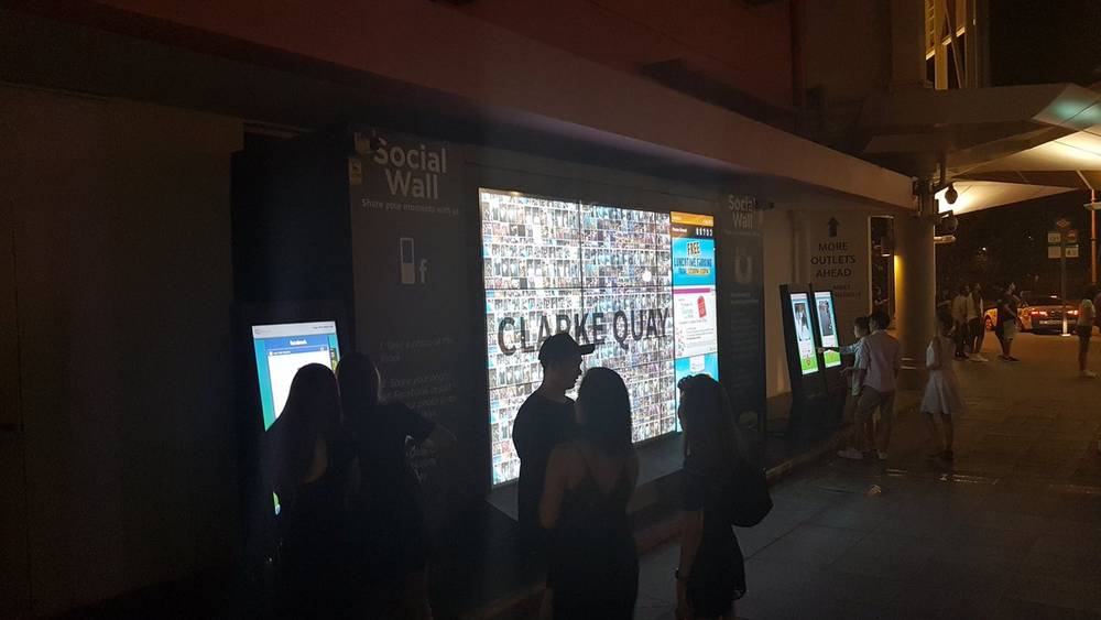 People looking at a large illuminated social media wall display at night, labeled Clarke Quay.