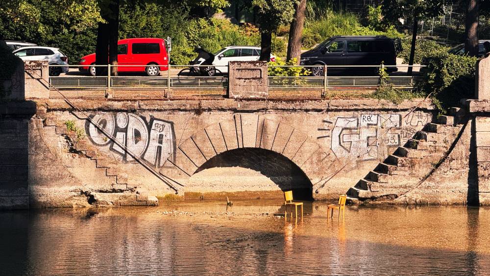 A river bank. The river is very low, there are two wooden chairs standing in it. The banking has a graffiti "oida" on it.