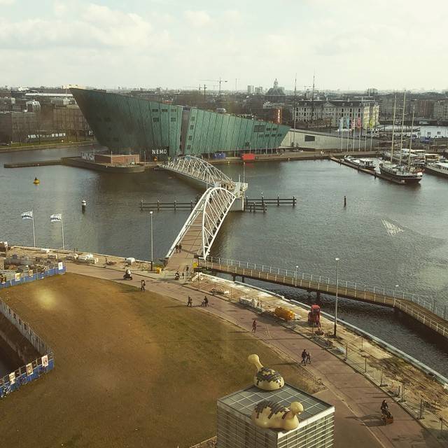 The green angular NEMO Science Museum building beside a harbor with a bridge.