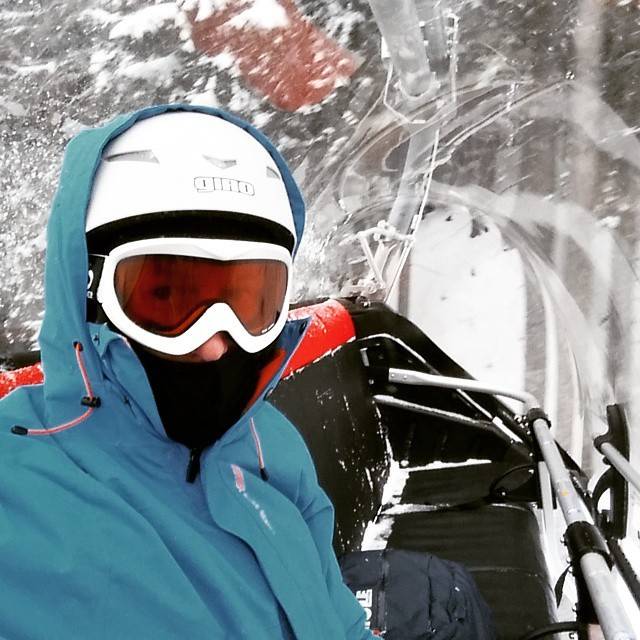 A snowboarder in a blue jacket and helmet rides a chairlift through falling snow.