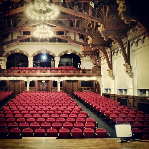 An ornate wood-paneled theater hall with rows of red seats and a chandelier.