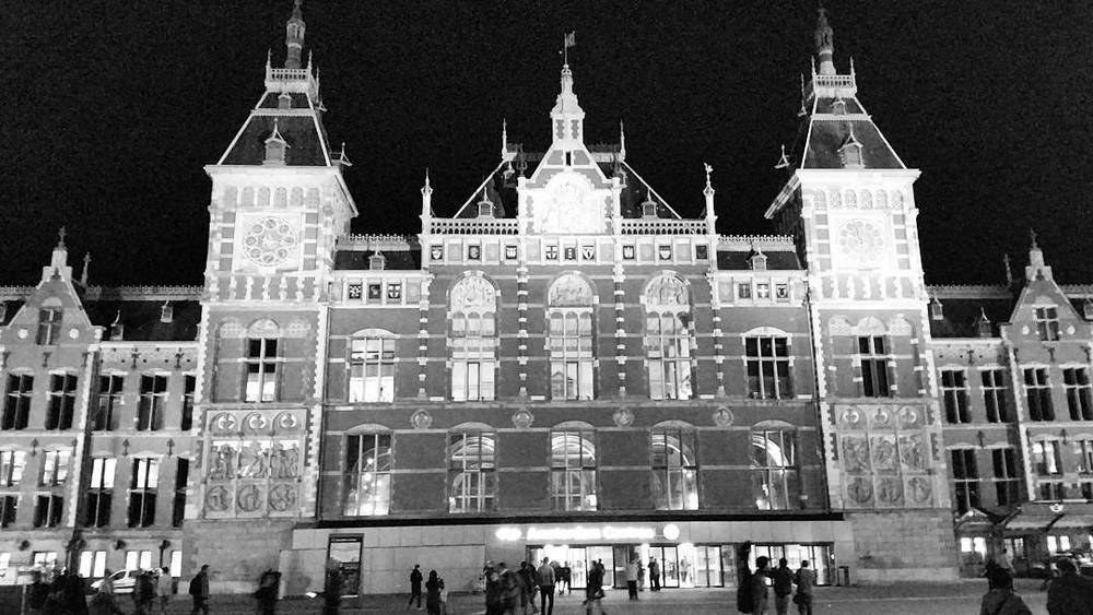 A black and white night view of the ornate facade of Amsterdam Centraal railway station, lit up.