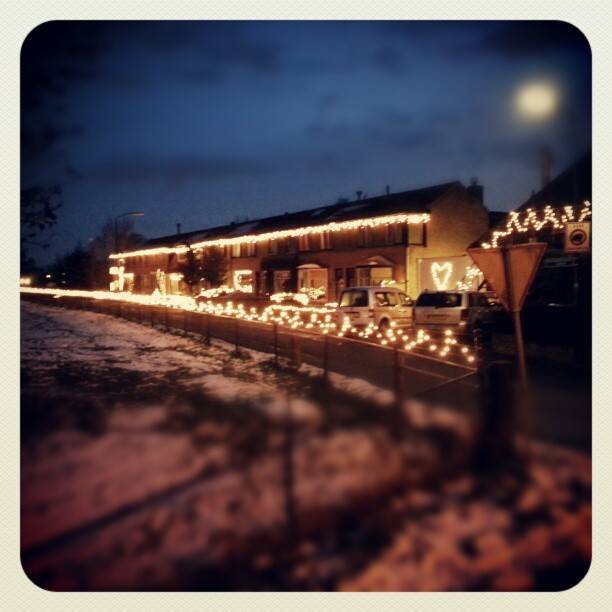 Rows of houses decorated with strings of Christmas lights, photographed at dusk in the snow.