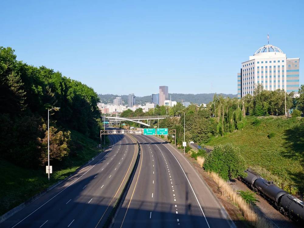 A multi-lane highway curves toward the downtown Portland, Oregon skyline, featuring modern office buildings and skyscrapers surrounded by lush green hills. The road is flanked by dense forests and includes green highway signs directing traffic toward the city center.