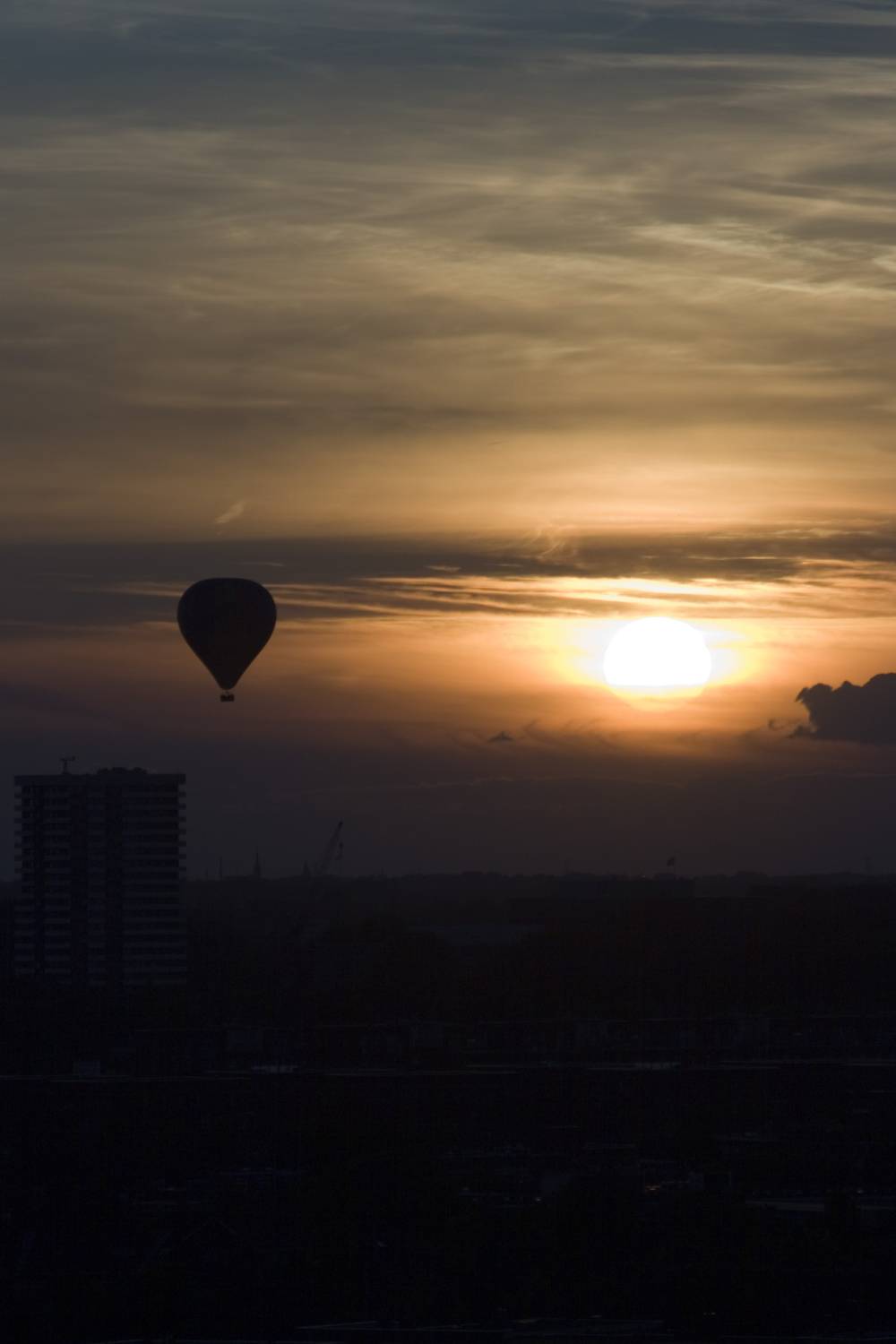 A hot air balloon drifting across a glowing orange sunset above a city skyline.