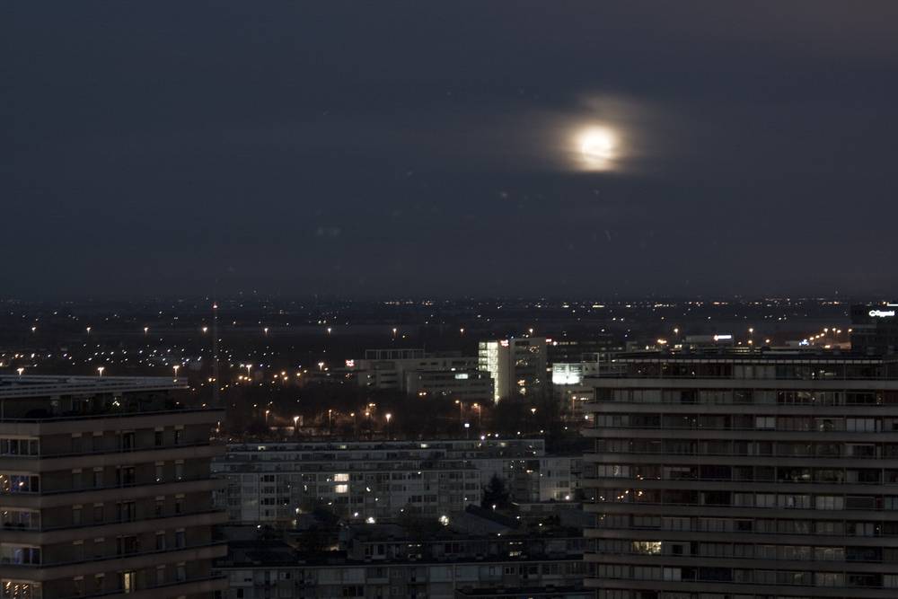 A pale moon sets behind apartment buildings in the early morning haze.