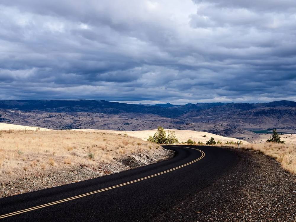 A two-lane asphalt road with double yellow center lines curves gently to the left and disappears over a ridge on Oregon State Highway 218 in Wheeler County, between the towns of Antelope and Fossil. Dry golden bunchgrass and scrubby high desert vegetation flank both sides of the road. A small cluster of juniper trees stands at the curve. Beyond the ridge, a vast panorama of rolling tan hills and eroded canyon country stretches to a distant mountain range under a dramatic, heavily overcast sky filled with layered blue-grey storm clouds. The overall palette is stark — dark asphalt, golden grassland, and brooding sky.