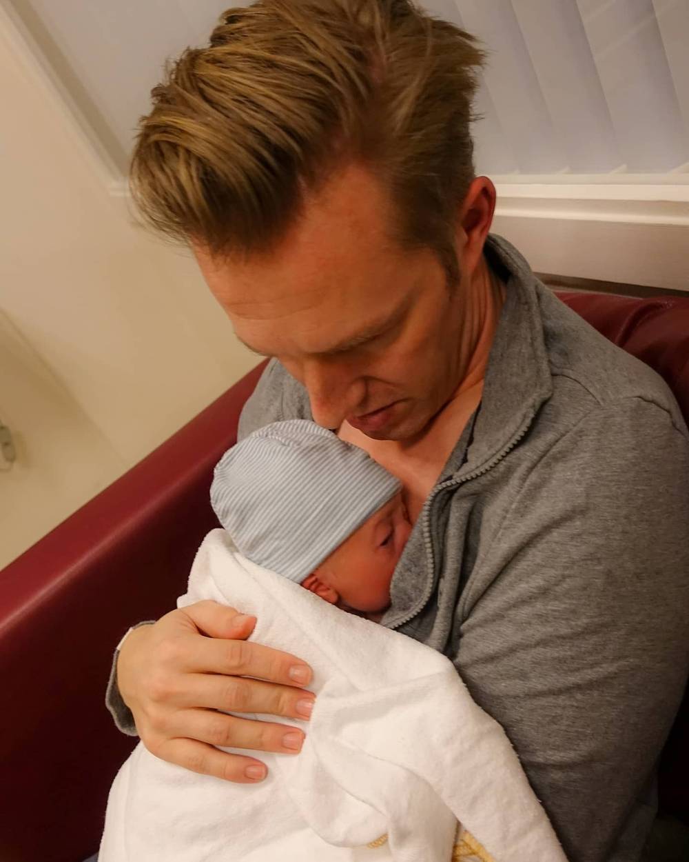 A man gently cradling a newborn wrapped in a white blanket and wearing a striped cap.