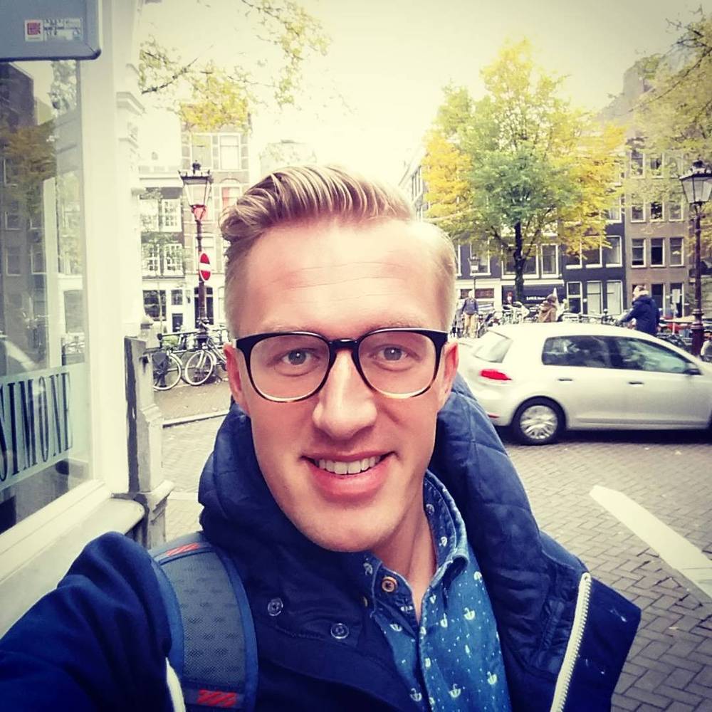 A selfie of a smiling man wearing glasses on an Amsterdam street with canal houses and bicycles.