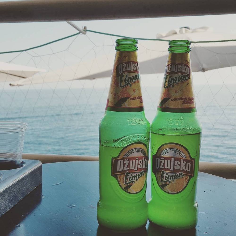 Two green glass bottles of Ožujsko Limun beer on an outdoor table overlooking the sea.