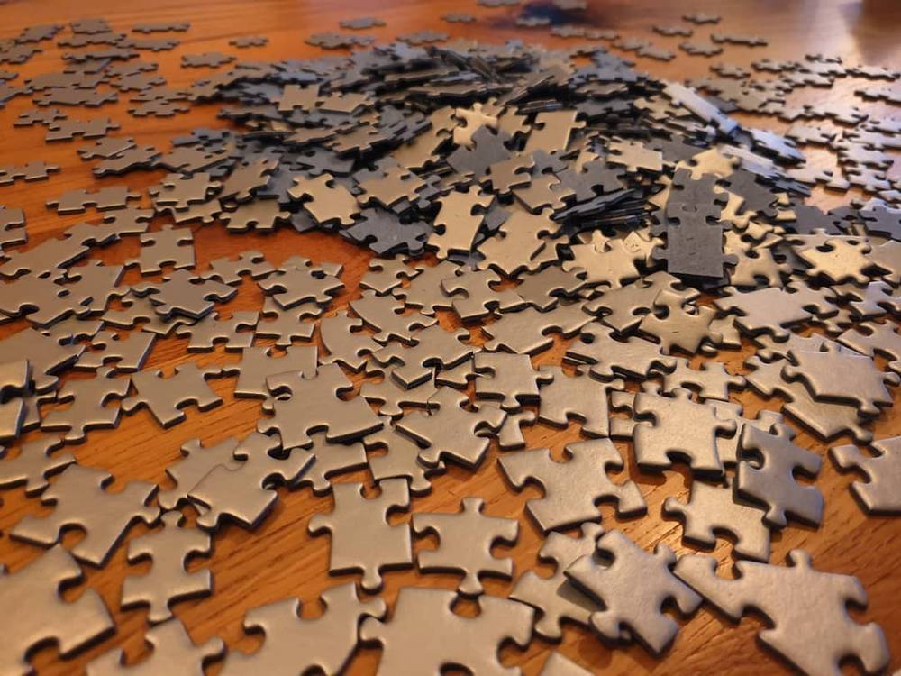 Hundreds of identical dark gray jigsaw puzzle pieces piled and scattered across a wooden table during sorting.