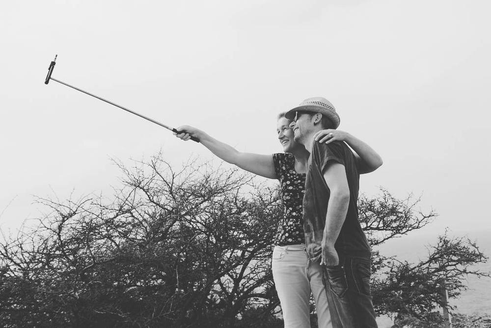 Black and white photo of a couple taking a selfie with a selfie stick near bushes.