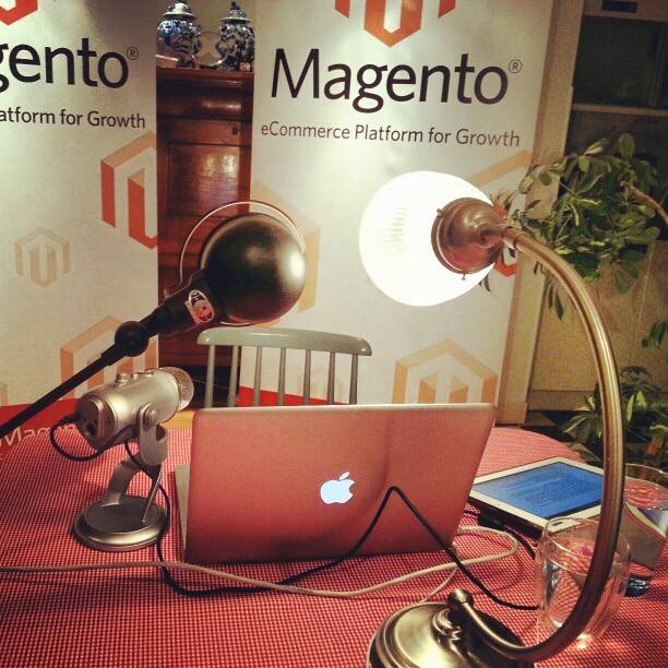 A laptop with an Apple logo set up on a red-and-white tablecloth at a Magento branded conference booth, with desk lamps and a photo of an antique cabinet on the backdrop.